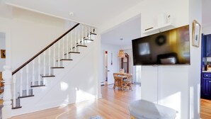 A modern living room with a staircase, a flat-screen TV mounted on a white wall, a wicker ottoman, and a blue patterned rug. - Cozy Historic Wakefield Home Close to Beaches (South Kingstown)