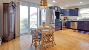 A modern kitchen with dark cabinets, a stainless steel refrigerator, and a dining area with wicker chairs and a glass table. - Cozy Historic Wakefield Home Close to Beaches (South Kingstown)