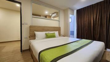 A hotel room with a bed, a wooden headboard, a green throw blanket, and a window with curtains.