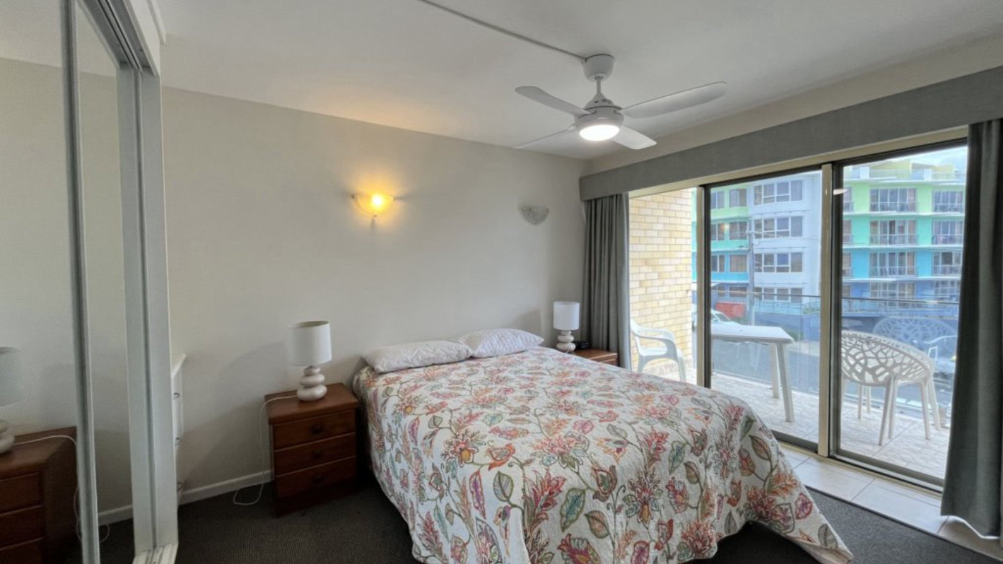 A bedroom with a bed, bedside tables, a ceiling fan, and a view of the outside.