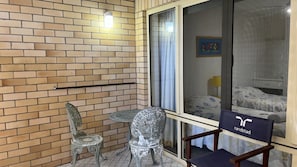 Un balcón con pared de ladrillo, dos sillas, una mesa y una cama visible a través de una ventana.
