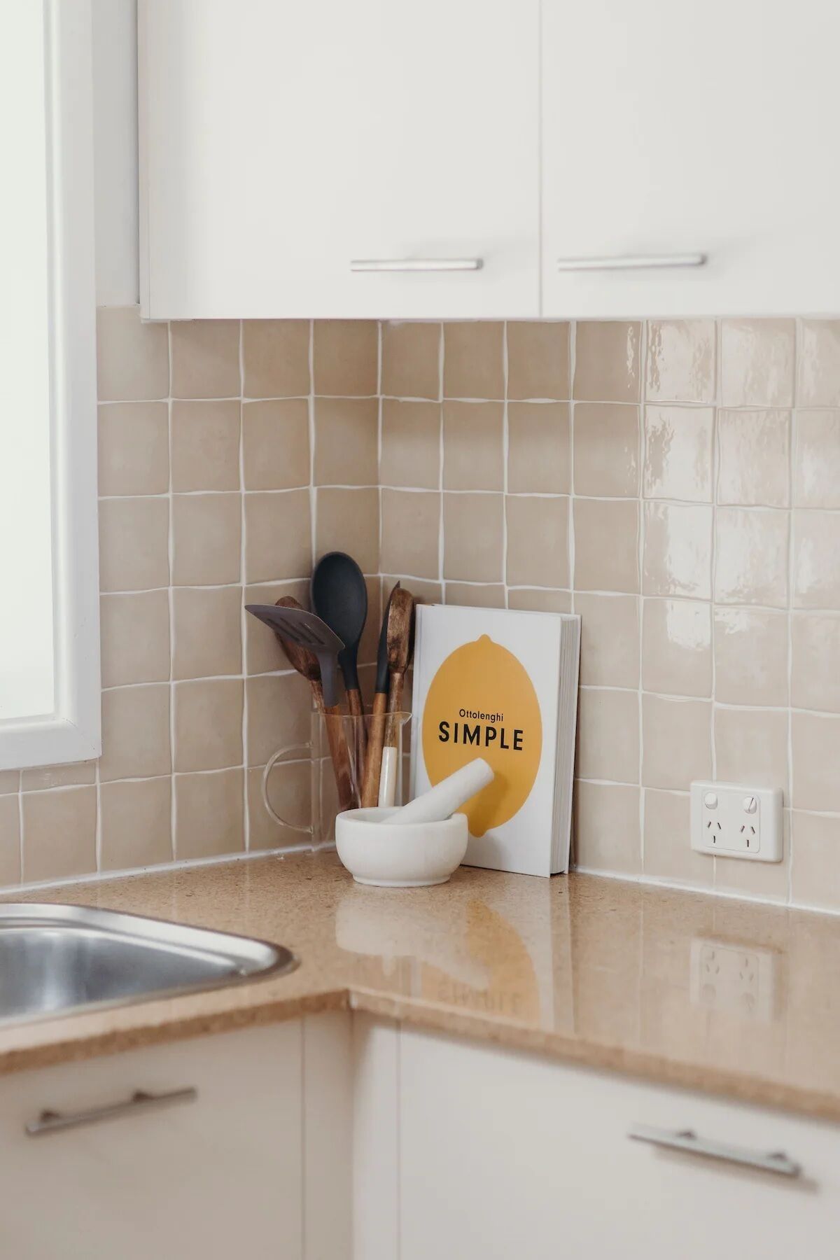 A kitchen with a tiled backsplash, a white cabinet, a countertop, a book titled 'Ottolenghi SIMPLE', and a set of wooden utensils.