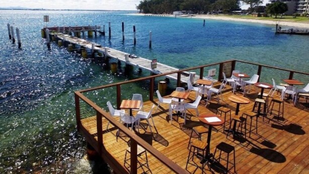 A wooden deck with tables and chairs overlooking a clear blue sea. - THE BAY. Effortless family fun. Pet friendly. (Nelson Bay)