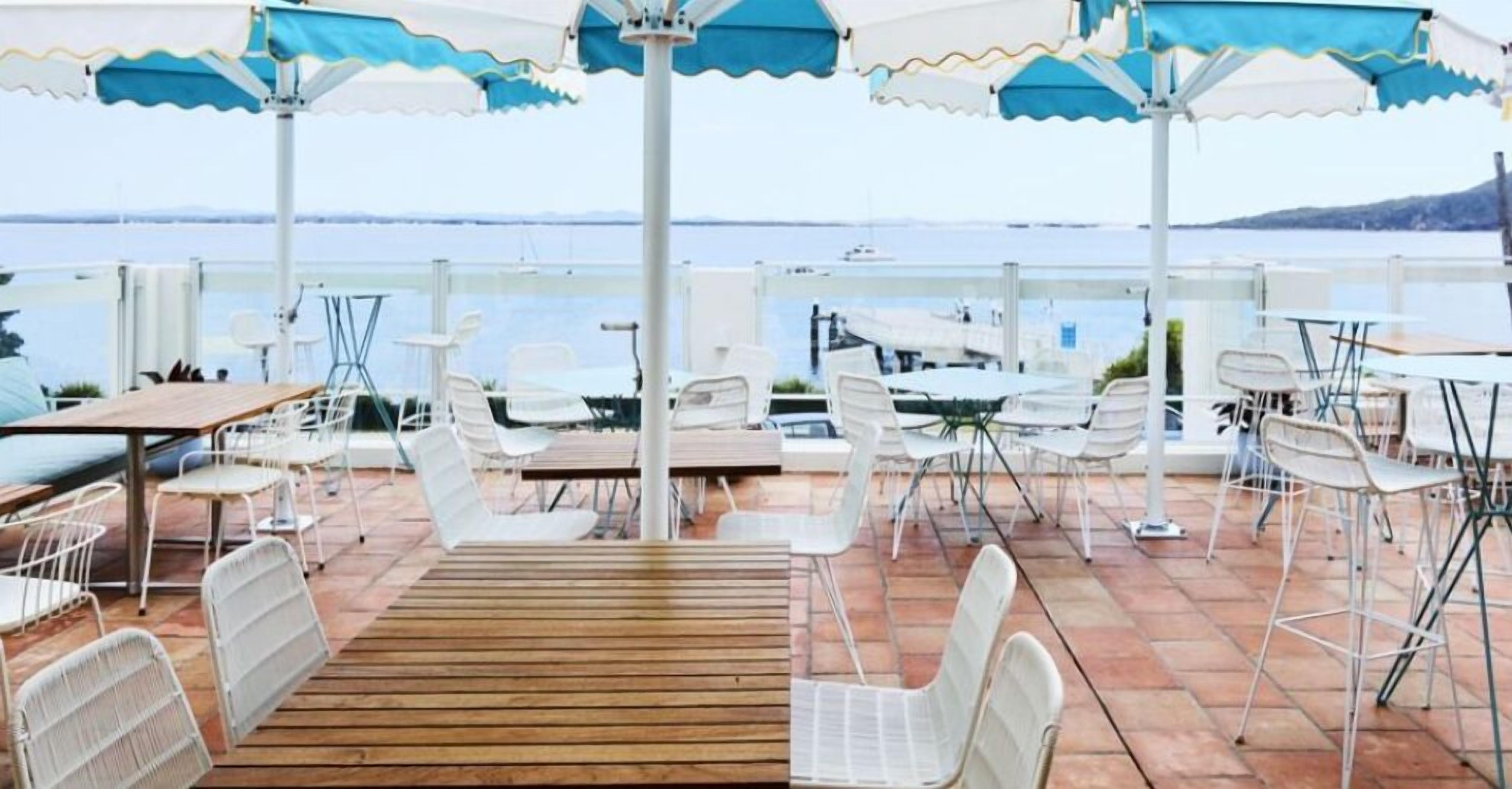 Patio esterno con sedie e tavoli in metallo bianco, ombrelloni blu e bianchi e vista sul mare.