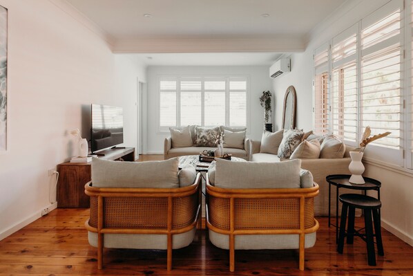 A living room with a sofa, two armchairs, a coffee table, a TV, and a window with blinds.