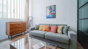 Ett modernt vardagsrum med en grå soffa, färgglada kuddar, en byrå i trä, ett soffbord i glas och en inramad bild av Paris.
