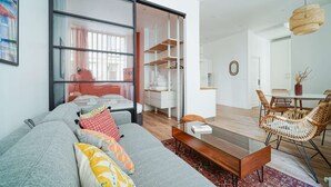 Ett modernt vardagsrum med en grå soffa, ett matbord i glas, stolar i rotting och ett stort fönster med utsikt över en stadspanorama.