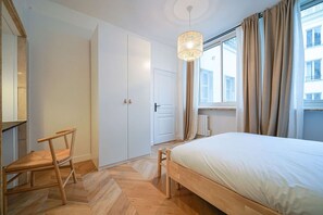 A bedroom with a bed, a chair, a wardrobe, a window with curtains, and a hanging light fixture.
