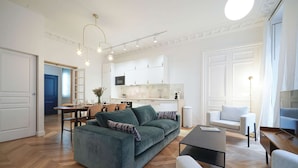 Un salon moderne avec un canapé vert, une table basse et un téléviseur. On y trouve également un coin repas avec une table et des chaises, et une cuisine à l’arrière-plan.