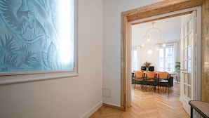 Un couloir doté d’un parquet en bois disposé en motif de chevron, une grande peinture abstraite sur le mur de gauche, et un coin repas avec une table et des chaises en bois, visibles à travers une porte ouverte.