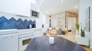 A modern kitchen with a dark countertop, white appliances, and a microwave above. There is a dining area with a dark table and a white candle. The room has a wooden floor and a sofa with cushions.