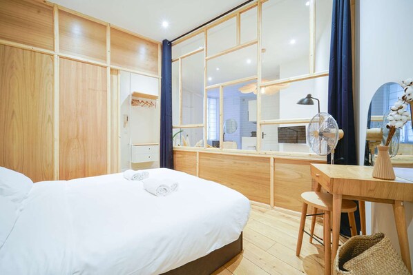 A modern hotel room with a large bed, a wooden desk, and a view through a glass partition.