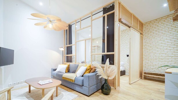 A modern living room with a grey sofa, wooden floor, and a large window with a view of a brick wall.