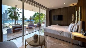 Een moderne hotelkamer met een groot bed, een glazen wand met uitzicht op zee en palmbomen, een flatscreen televisie en een klein tafeltje.