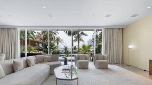 Een moderne woonkamer met een ruime hoekbank, twee fauteuils en een glazen salontafel. De kamer heeft grote ramen met gordijnen, met uitzicht op palmbomen en een stadsbeeld.