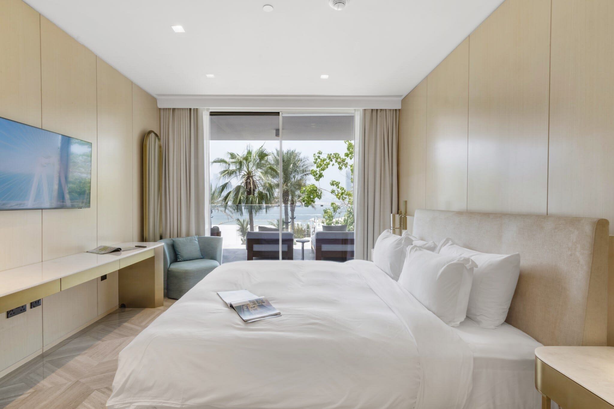 A hotel room with a large bed, a desk, a TV, and a view of palm trees and the sea.