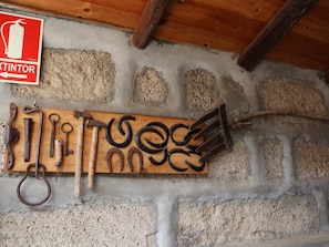 A rustic interior with a wooden tool rack against a stone wall, a fire extinguisher sign, and a wooden beam ceiling.