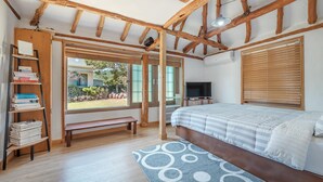 A bedroom with a wooden ceiling, a bed with a striped duvet, a bookshelf, a television, and a window with blinds. - Jeju Motungedoljib Dogchaepension (Jeju City)