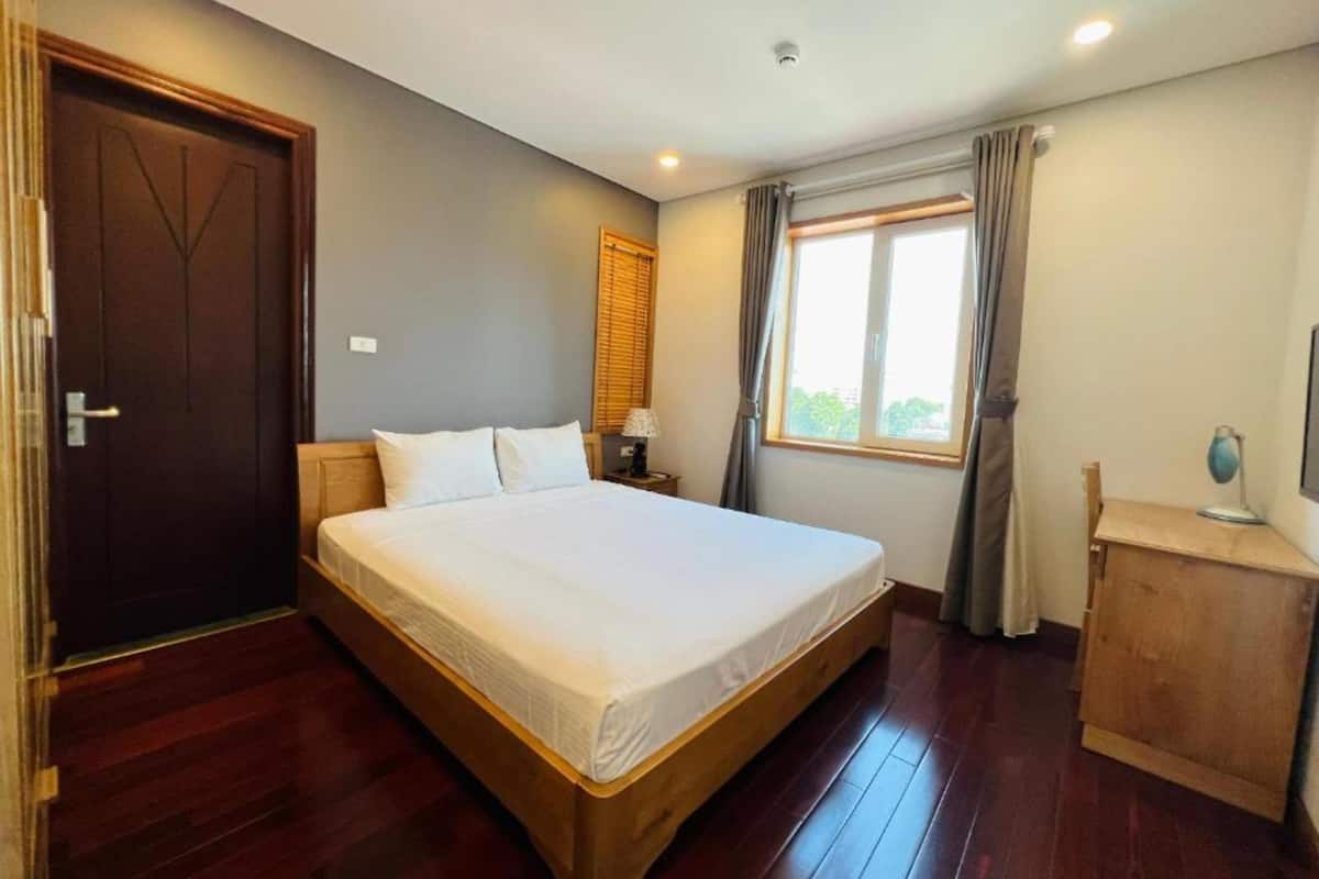 A bedroom with a wooden bed, a nightstand, a desk, a window with curtains, and a door.