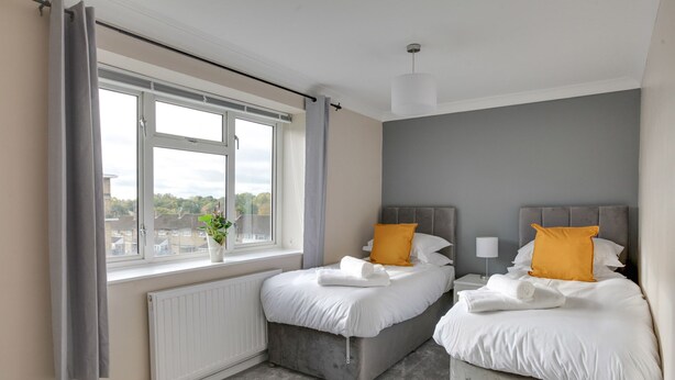A bedroom with two beds, a window with curtains, a radiator, and a plant. - BookedUK - Bright flat in Harlow (Harlow)