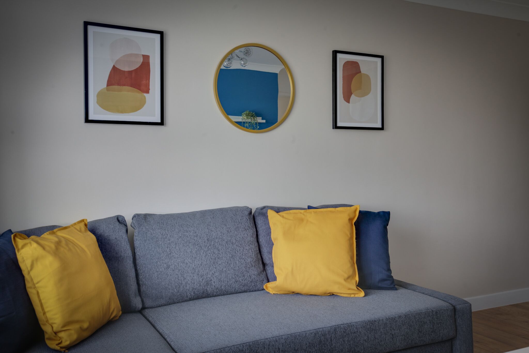 A grey sofa with yellow and blue cushions, a round mirror, and two framed abstract paintings on the wall.