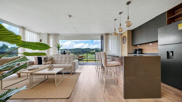 A modern living room with a sofa, coffee table, and bar stools. There is a kitchen area with a refrigerator and a dining table. Large windows provide a view of the outdoors. - Olive 301 · Amazing 2BR Luxury Apt in Alto Las Palmas (Alto Palmas)