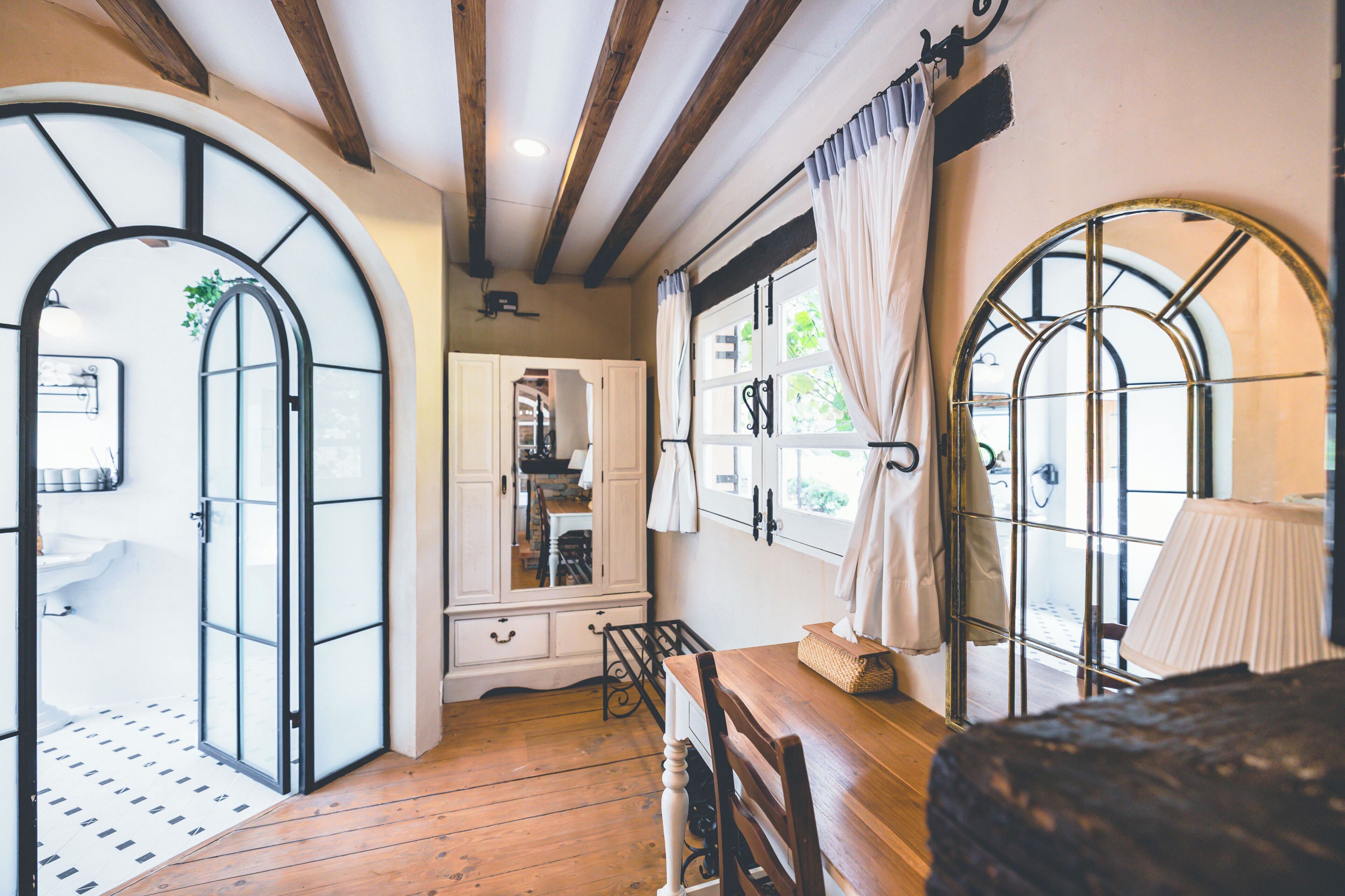 A room with wooden flooring, a wooden table, a chair, a window with curtains, and a mirror.