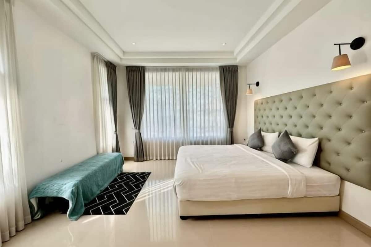 Une chambre à coucher moderne avec un grand lit, un banc, une tête de lit capitonnée et une fenêtre avec des rideaux.