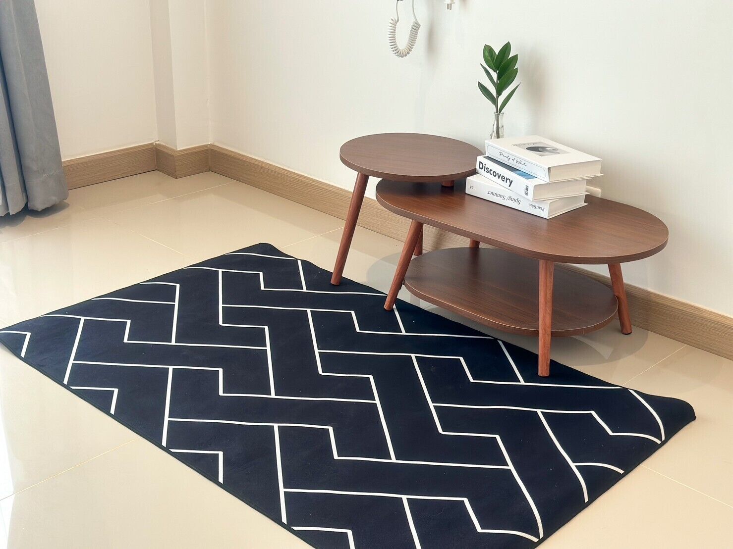 A room with a black and white chevron-patterned rug, a small round wooden table with a plant and books, and a plain white wall.