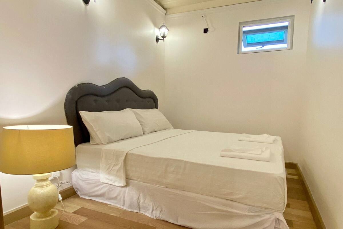 Une chambre à coucher avec un lit, une lampe, un luminaire fixé au mur et une petite fenêtre.