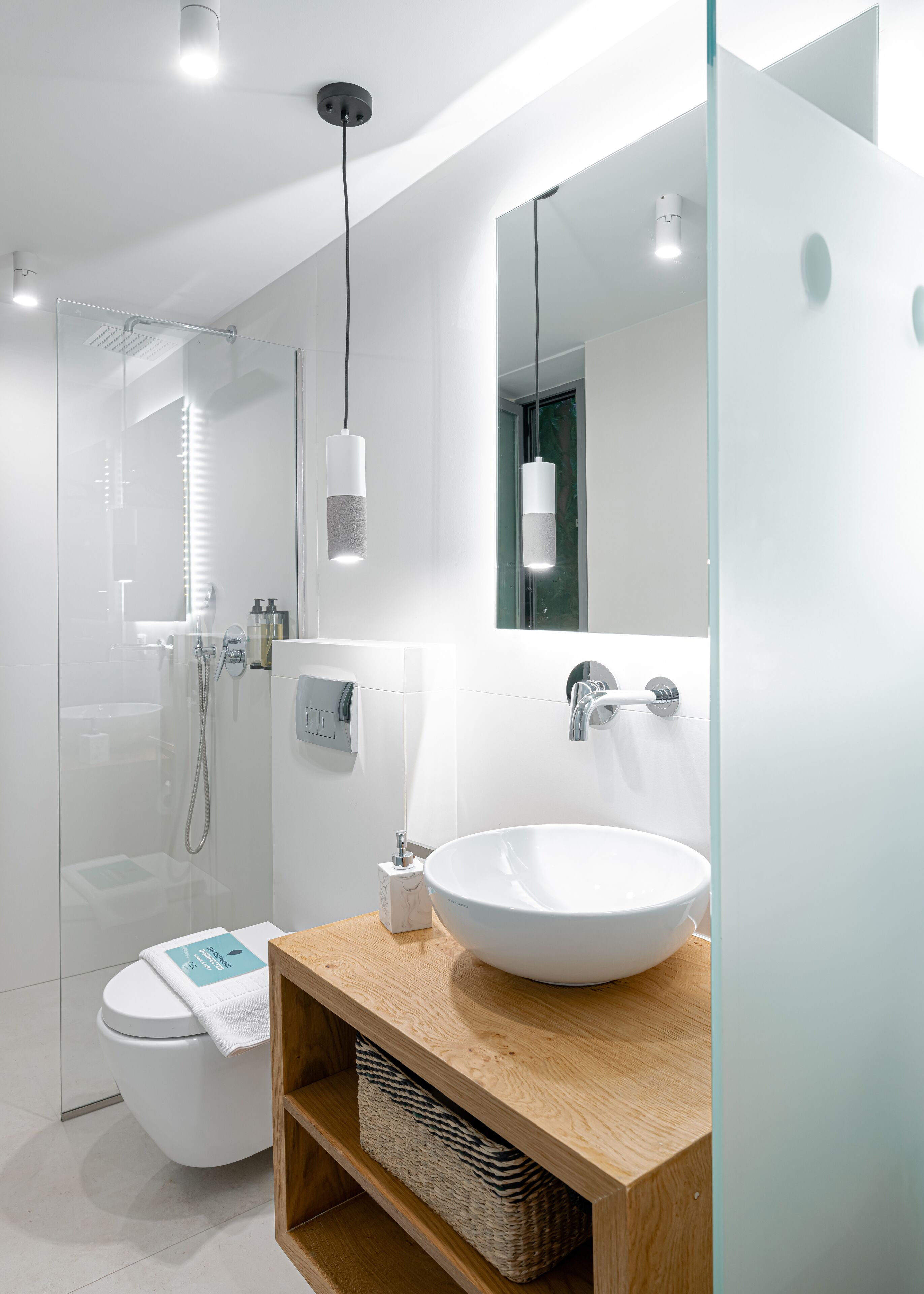 A modern bathroom with a large white sink, a wooden vanity, a glass shower enclosure, and a mirror with a light fixture above it.