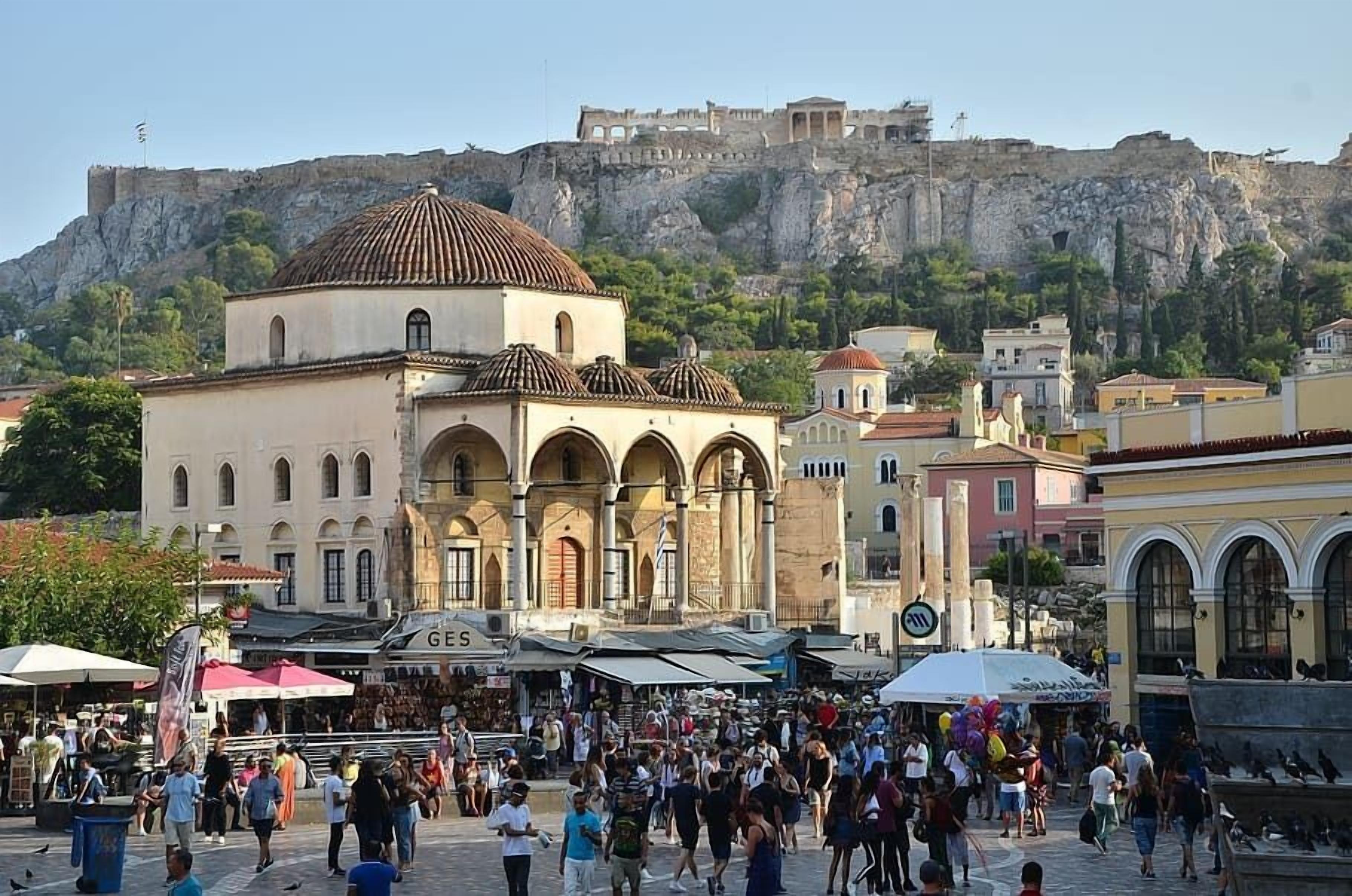 A bustling square with a prominent historical building featuring arches and a dome, surrounded by modern structures and a cliff with ruins in the background.