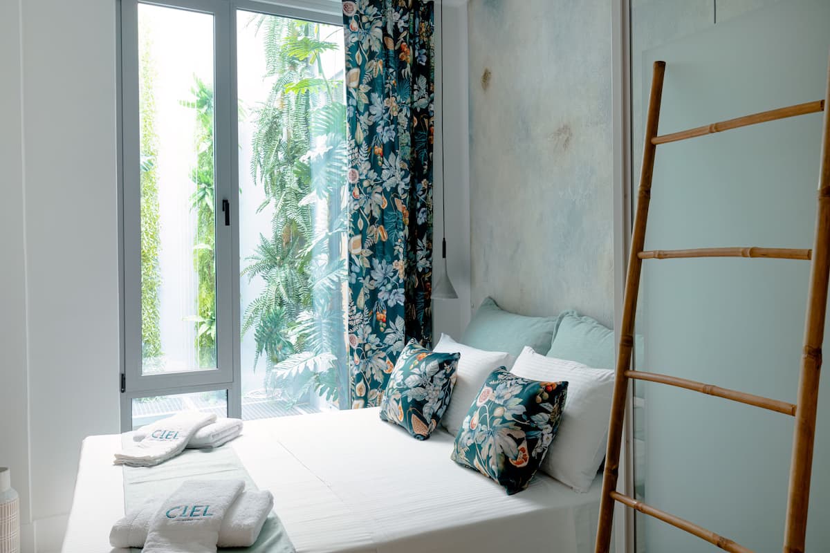 Quarto com cama, cortinas florais e vista para o verde lá fora.