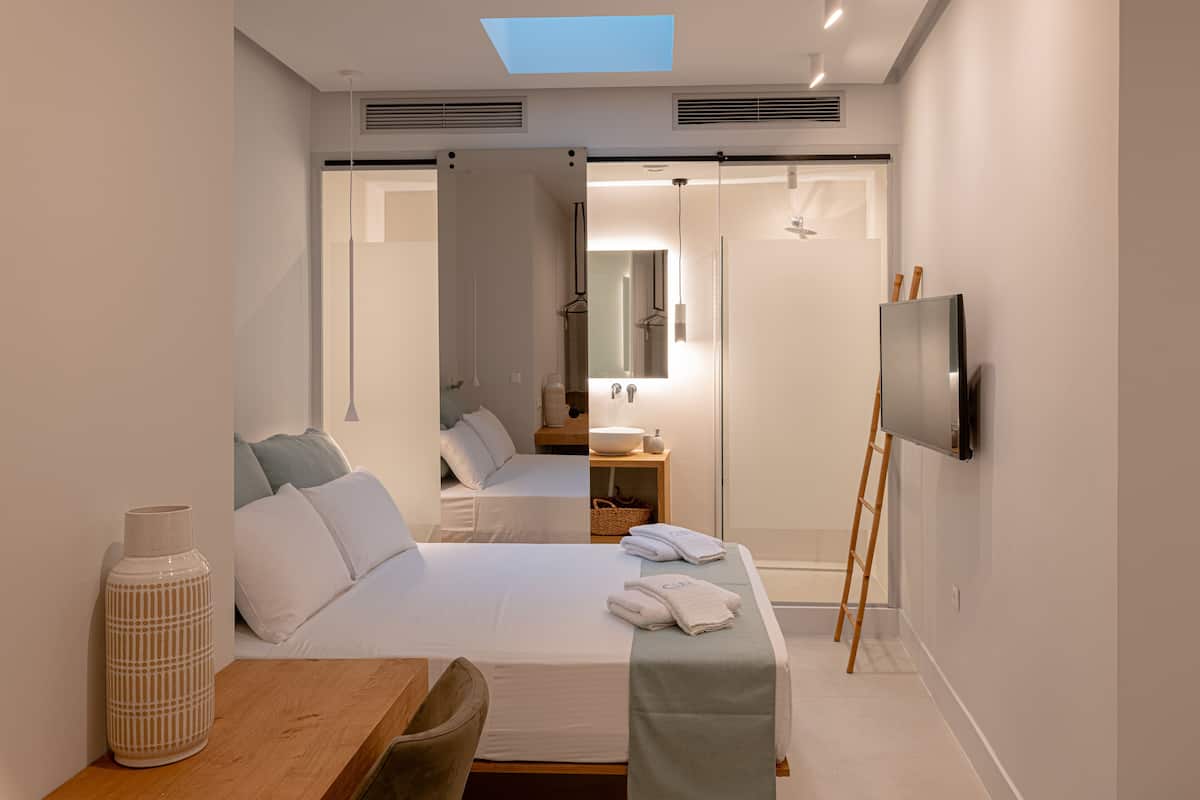 Quarto de hotel moderno com cama, escrivaninha, televisão e banheiro visível através de uma porta de vidro.