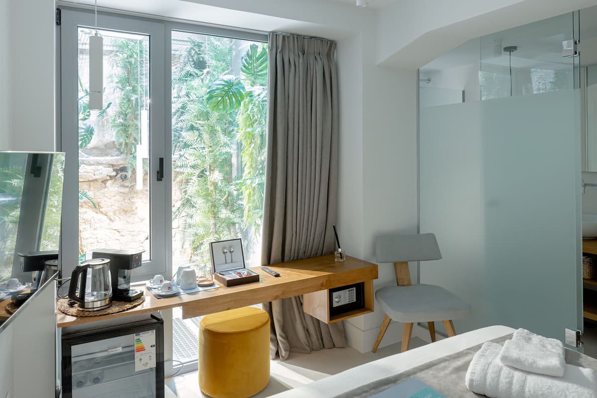 Quarto de hotel com uma cama, uma escrivaninha com computador, cafeteira e vista para o verde lá fora.