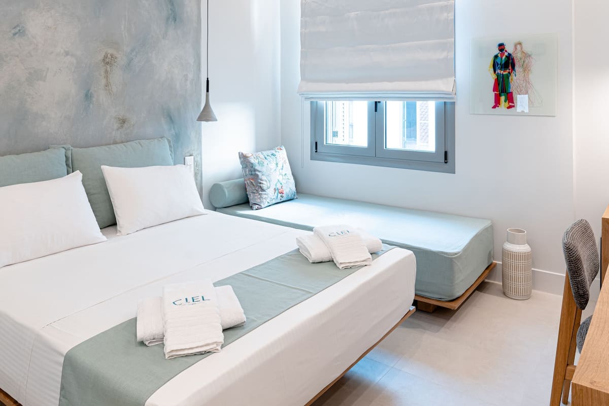 Quarto moderno com uma cama grande, um banquinho, janela com persianas, uma mesinha e uma prateleira na parede.