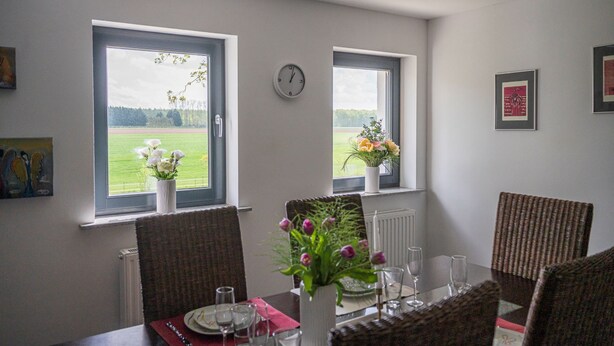 Un coin repas avec deux fenêtres, une horloge et une table dressée pour deux personnes, avec des chaises.