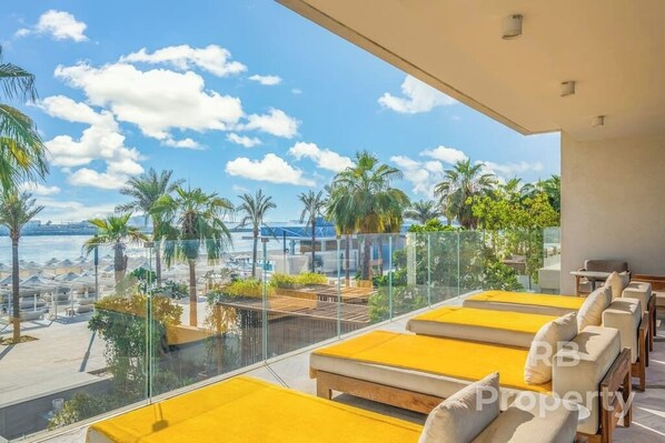 A balcony with yellow cushions, a glass railing, and a view of palm trees and a body of water.