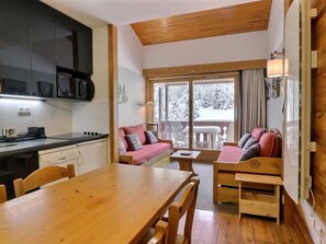 A cozy cabin interior with a wooden dining table, a sofa with cushions, a kitchen area with cabinets and a stove, and a large window with a view of snow-covered trees.