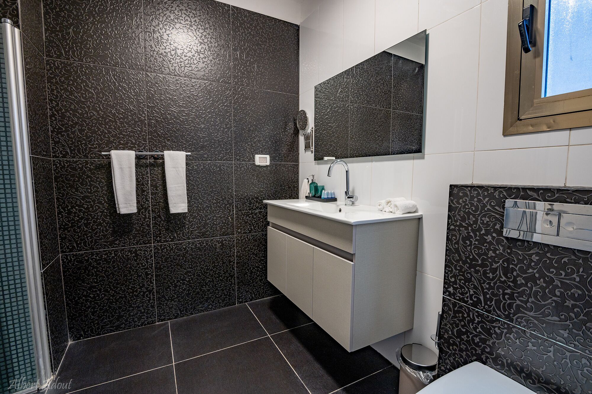 Une salle de bain moderne avec des carreaux à motifs foncés, un meuble-lavabo blanc avec un miroir et un espace douche.