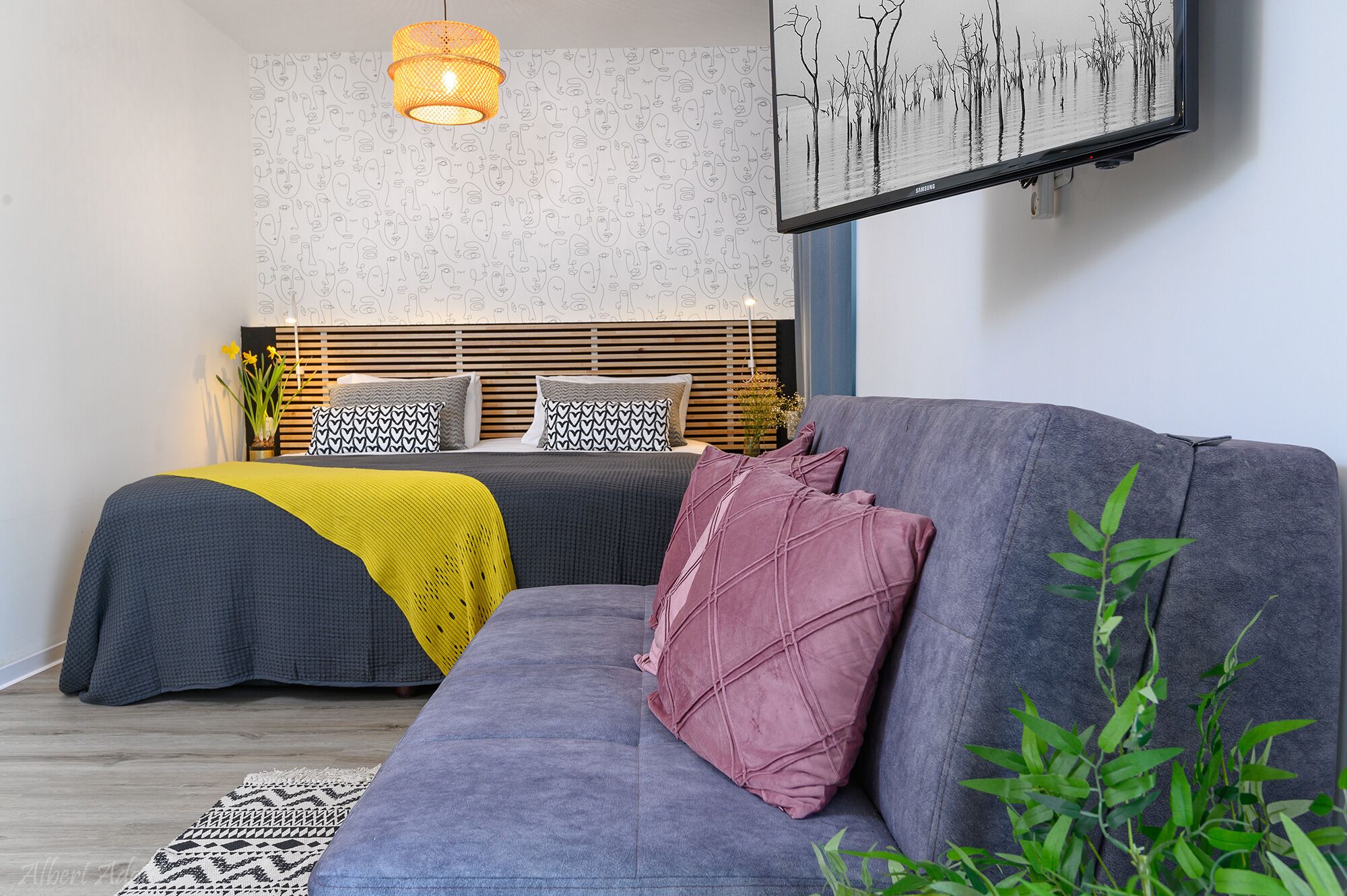 A modern bedroom with a grey sofa, a bed with patterned pillows, a yellow throw, and a wall-mounted TV.