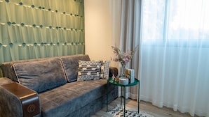 A living room with a brown sofa, patterned pillows, a small green side table with a vase of flowers, and sheer curtains. - Kamaya Suites (Emek HaYarden)