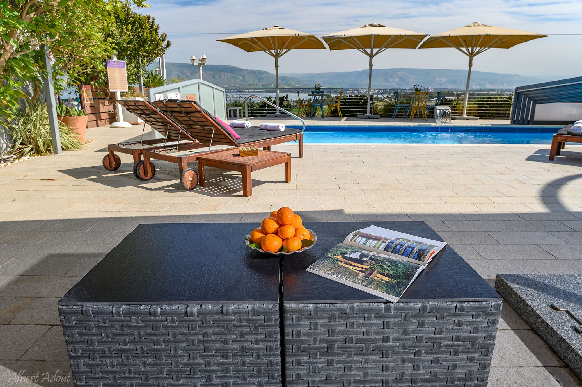 A poolside area with lounge chairs, umbrellas, and a table with a bowl of oranges and a magazine.