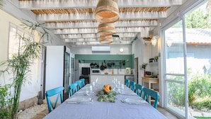 A dining area with a long table set for six, blue chairs, a hanging woven light fixture, and a view of the kitchen and outdoors through large windows. - Kamaya Suites (Emek HaYarden)