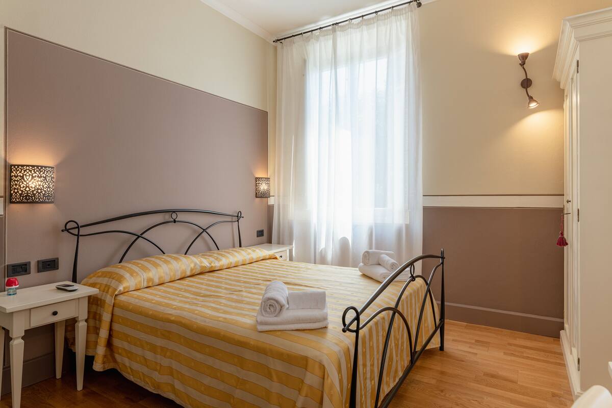 A neatly made bed with striped bedding, a bedside table with a lamp, and a window with sheer curtains.