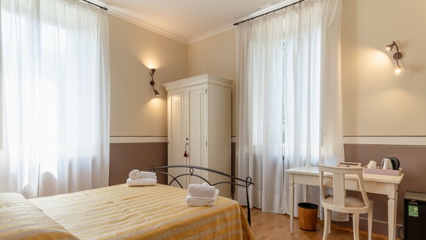 A hotel room with a bed, a desk, a chair, a wardrobe, and a trash bin. - Villa Romantica (Lucca)