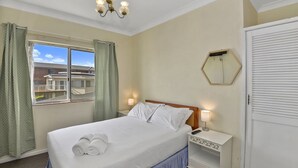 A hotel room with a bed, a nightstand, a lamp, a window with curtains, and a view of buildings outside. - Beachside at The Entrance (The Entrance)