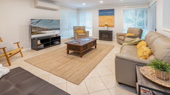 A living room with a sofa, armchair, coffee table, TV, and a fireplace. - Frangipani Beach House (Boomerang Beach)