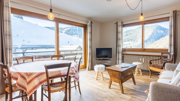 A room with a dining table, chairs, a sofa, a TV, and large windows with a view of a snowy landscape. - 3* Apartment, 2 bedrooms, 6 guests, near slopes, WIFI (La Clusaz)