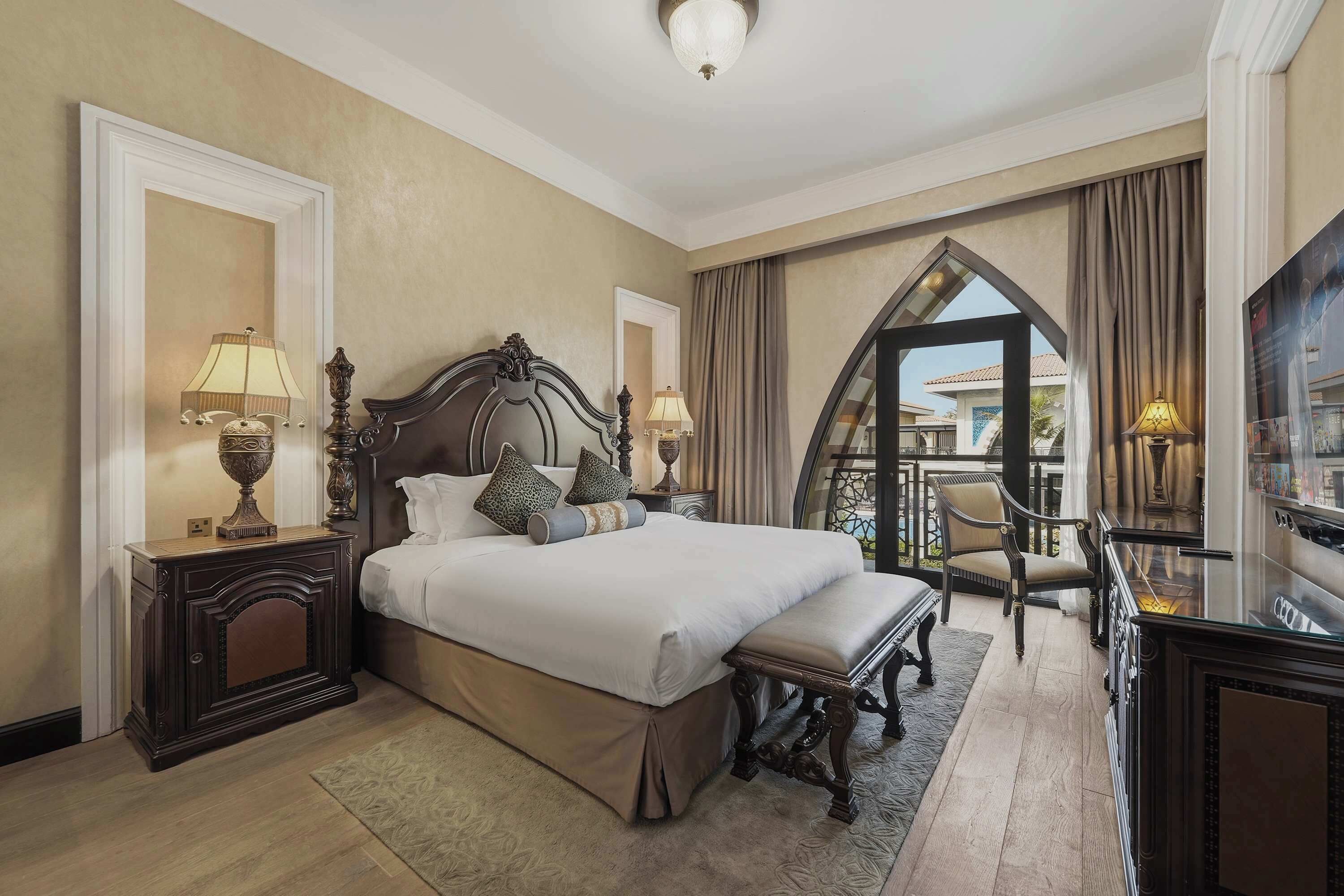 Quarto de hotel com uma cama grande, duas mesas de cabeceira com luminárias, uma cadeira, uma escrivaninha e vista para o exterior.
